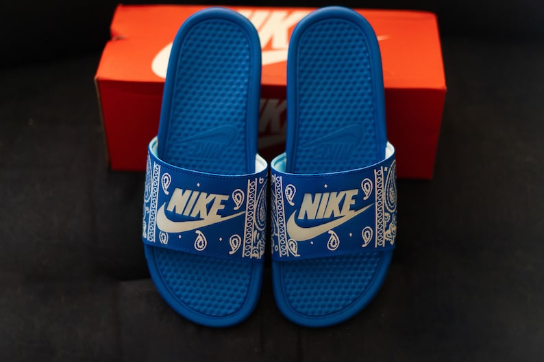 May include: Pair of blue Nike slides with white paisley pattern straps and the Nike logo. The slides are on a black surface with a red Nike box in the background.