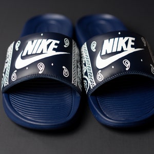 May include: A pair of navy blue Nike slides. The slides feature a white Nike logo on the strap, which is decorated with a white paisley pattern. The footbed is navy blue with a textured design.