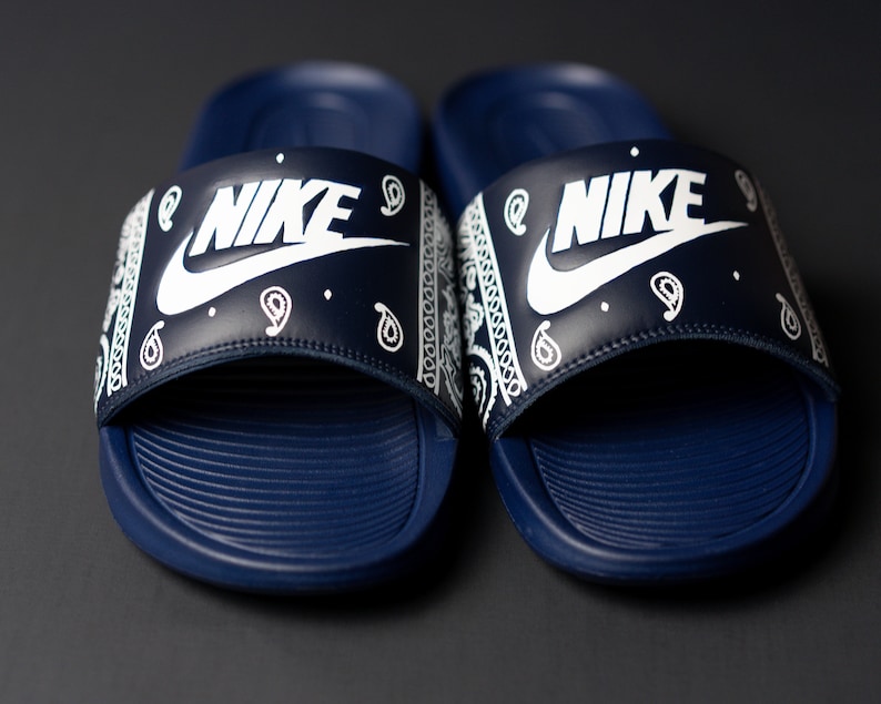 May include: A pair of navy blue Nike slides. The slides feature a white Nike logo on the strap, which is decorated with a white paisley pattern. The footbed is navy blue with a textured design.
