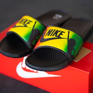K&ouml;nnte beinhalten: Schwarze Nike-Slides mit gelbem, gr&uuml;nem und rotem Design. Die Slides zeigen das Nike-Logo in Schwarz und eine Silhouette einer Person. Die Slides liegen auf einer roten Nike-Box.