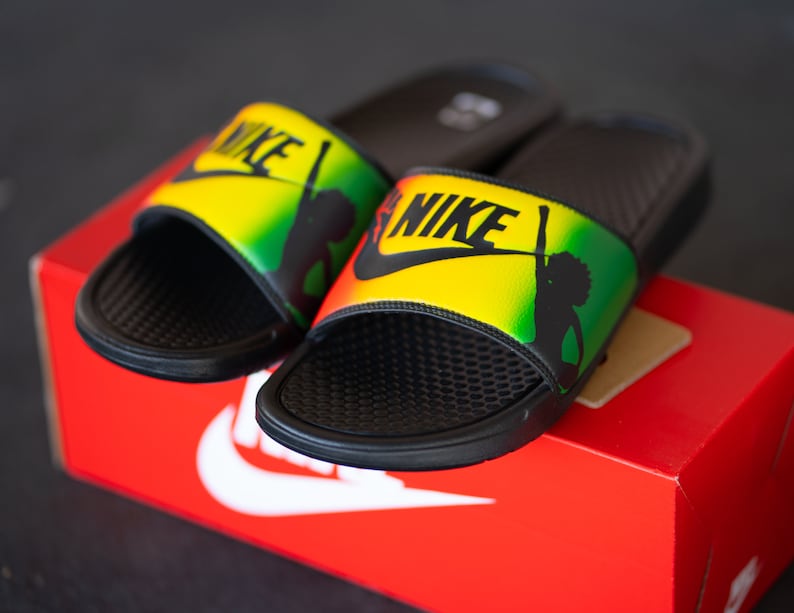 K&ouml;nnte beinhalten: Schwarze Nike-Slides mit gelbem, gr&uuml;nem und rotem Design. Die Slides zeigen das Nike-Logo in Schwarz und eine Silhouette einer Person. Die Slides liegen auf einer roten Nike-Box.