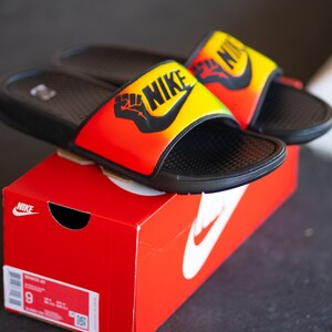 K&ouml;nnte beinhalten: Schwarze Nike-Slides mit strukturiertem Fu&szlig;bett, auf einer roten Nike-Box. Die Slides haben ein gelb-orange-rotes Farbverlauf-Design, das Nike-Logo und eine schwarze Faust-Grafik.