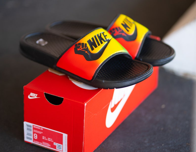 K&ouml;nnte beinhalten: Schwarze Nike-Slides mit strukturiertem Fu&szlig;bett, auf einer roten Nike-Box. Die Slides haben ein gelb-orange-rotes Farbverlauf-Design, das Nike-Logo und eine schwarze Faust-Grafik.
