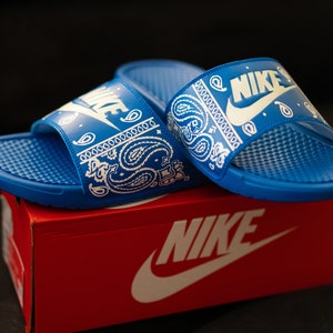 May include: Blue Nike slides with white paisley pattern and Nike logo. The slides are on top of a red Nike box with the Nike logo in white. The slides have a textured footbed.