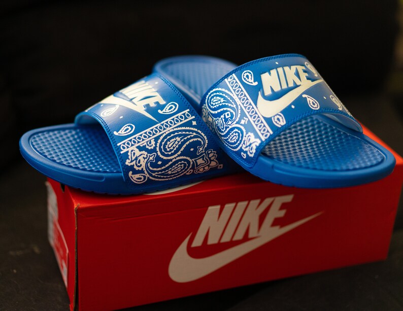 May include: Blue Nike slides with white paisley pattern and Nike logo. The slides are on top of a red Nike box with the Nike logo in white. The slides have a textured footbed.