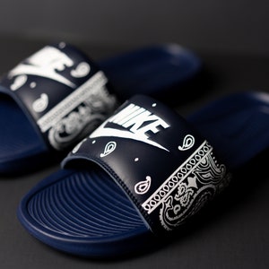 May include: A pair of navy blue Nike slides with white paisley print on the strap. The Nike logo is prominently displayed in white. The slides have a textured footbed and are set against a dark background.