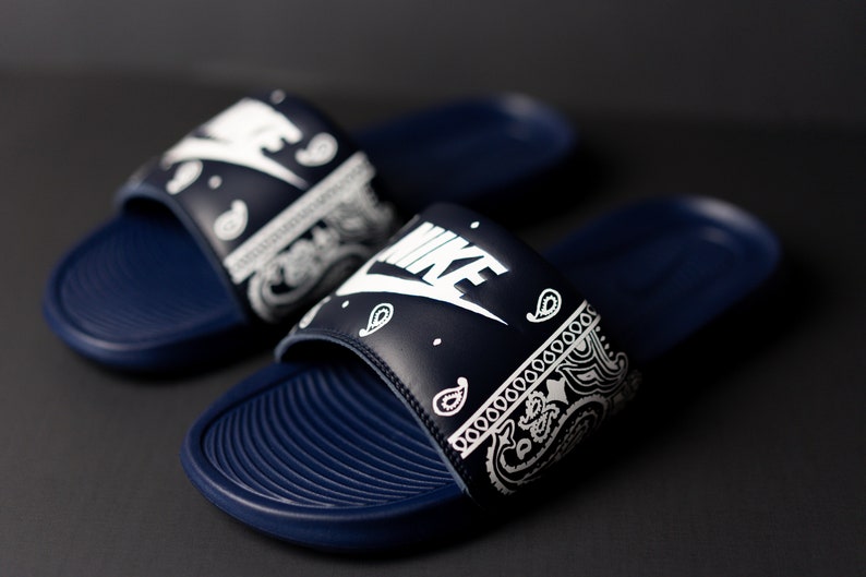 May include: A pair of navy blue Nike slides with white paisley print on the strap. The Nike logo is prominently displayed in white. The slides have a textured footbed and are set against a dark background.