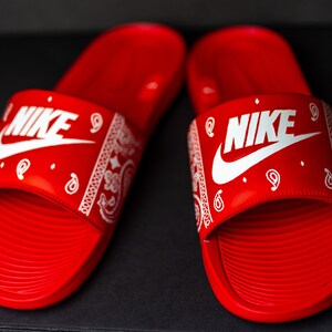 May include: Pair of red Nike slides with white Nike logo and white paisley pattern on the strap. The slides are on a black surface. The slides are a type of footwear.