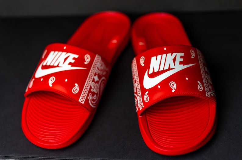 May include: Pair of red Nike slides with white Nike logo and white paisley pattern on the strap. The slides are on a black surface. The slides are a type of footwear.
