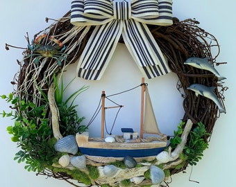 May include: A nautical-themed wreath with a grapevine base, featuring a blue and white striped bow, a miniature sailboat, seashells, and two dolphin figurines. A blue crab and greenery add to the coastal design.