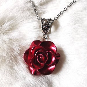 Könnte beinhalten: Eine silberfarbene Halskette mit einem Anhänger in Form einer roten Rose. Der Rosenanhänger hat detaillierte Blütenblätter und ein metallisches Finish. Die Kette ist filigran, und der Anhänger hängt an einem verzierten Bügel.