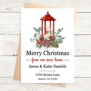 Pode incluir: Uma lanterna vermelha com uma vela branca dentro, cercada por vegetação e bagas vermelhas. O texto "Feliz Natal da nossa nova casa" está escrito acima dos nomes "Jason & Katie Daniels". O endereço "1234 Spruce Lane, Denver, CO 12345" está impresso abaixo dos nomes.