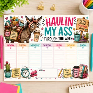 Op de afbeelding: Een weekplanner bureaulegger met een ezelillustratie, de tekst "HAULIN' MY ASS THROUGH THE WEEK" en "IT'S FINE. I'M FINE. EVERYTHING IS FINE." De lay-out bevat zeven dagkolommen, een notitiesectie en kleurrijke afbeeldingen van koffie, planten en kantoorbenodigdheden.