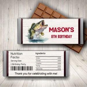 Puede incluir: Un envoltorio de barra de chocolate con temática de pesca. El envoltorio es blanco con un patrón de veta de madera marrón. Presenta una lubina de dibujos animados saltando fuera del agua. El texto "MASON'S 8TH BIRTHDAY" está impreso en rojo en el envoltorio. La parte posterior del envoltorio tiene una etiqueta de información nutricional y una lista de ingredientes.