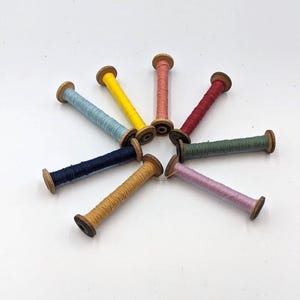 May include: A collection of colorful thread spools arranged in a circular pattern. The spools are made of wood and filled with thread in various colors, including blue, yellow, pink, and green. The spools are approximately 1 inch in length.