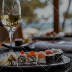 K&ouml;nnte beinhalten: Ein Teller mit Sushi-Rollen mit verschiedenen F&uuml;llungen, darunter Lachs, Avocado und Gurke. Die Rollen sind in einer Reihe auf einem schwarzen Teller angeordnet. Im Hintergrund steht ein Glas Wei&szlig;wein.