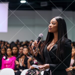 May include: A woman in a black blazer speaks into a microphone while addressing a seated audience. She is wearing a necklace and has long black hair.