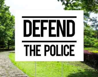 Pode incluir: Uma placa branca com as palavras "DEFEND THE POLICE" em letras pretas em negrito. A placa está em estacas de metal e está colocada em uma área gramada com árvores ao fundo. O texto "FREE SPEECH SIGNS" está na parte inferior.