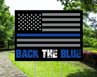 Pode incluir: Uma placa de jardim preta com um design estilizado da bandeira americana, com uma linha azul, listras cinzas e estrelas brancas. As palavras "BACK THE BLUE" estão em azul e branco. A placa é montada em duas estacas de metal.