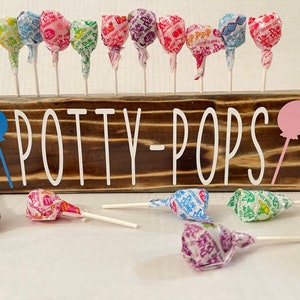 May include: A wooden sign with the text "POTTY-POPS" painted in white. The sign has a blue balloon on the left and a pink balloon on the right. There are 14 colorful lollipops in the background.