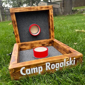 May include: A wooden cornhole game with a dark stain and the text "Camp Rogalski" in white. The game has a red target and a red cylinder. The interior is lined with gray felt.