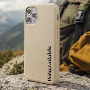 以下が含まれることがあります： ベージュ色の携帯電話ケース。黒字で「Biodegradable」の文字が縦にプリントされています。ケースはテクスチャ加工が施されており、カメラ用の切り抜きがあります。背景には山岳風景とバックパックが写っています。