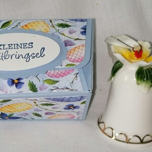 Könnte beinhalten: Eine kleine Geschenkbox mit floralem Muster und dem deutschen Text "Kleines Mitbringsel" neben einer weißen Keramikglocke mit gelber Blume und goldenem Rand. Die Box ist hellblau.