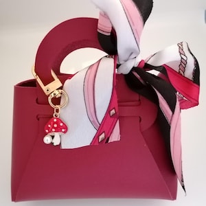 Könnte beinhalten: Burgunderfarbene Handtasche mit goldfarbenem Verschluss und einem Pilz-Anhänger. Ein gemustertes Tuch in Weiß, Rosa und Schwarz ist am Griff gebunden. Der Pilz-Anhänger hat einen weißen Stiel und einen roten Hut mit weißen Punkten.