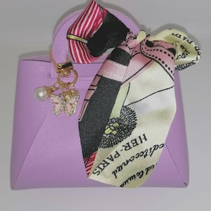 Könnte beinhalten: Eine kleine, lila Handtasche mit einem dekorativen Schal und einem goldfarbenen Anhänger. Der Schal hat ein Blumen- und Textdesign mit rosa, schwarzen und gelben Akzenten. Der Anhänger enthält eine Perle und einen rosa Schmetterling.