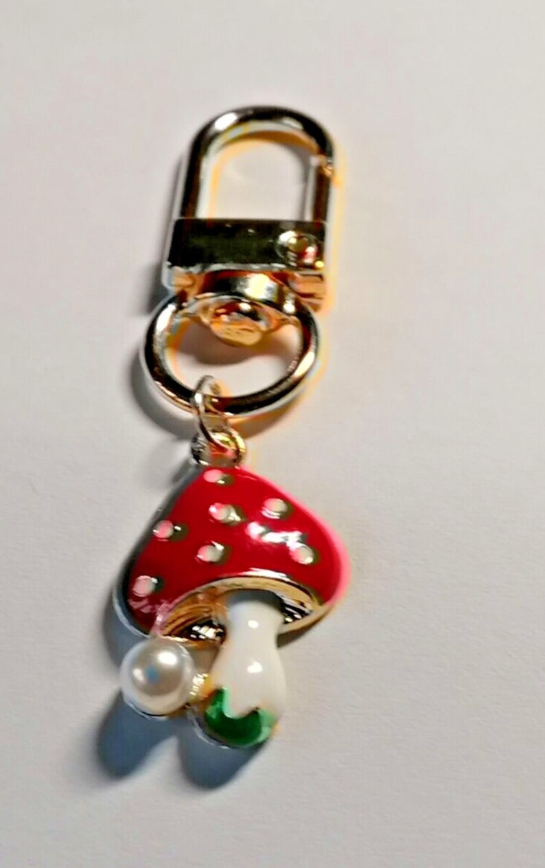 Op de afbeelding: Een goudkleurige sleutelhanger met een rode paddenstoelbedel. De paddenstoel heeft witte stippen, een witte steel en een groene basis. Een imitatieparel is bevestigd. De sleutelhanger heeft een karabijnhaak.