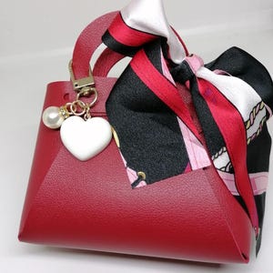 Op de afbeelding: Een kleine, rode handtas met een handvat aan de bovenkant en een decoratieve sjaal die aan de bovenkant is vastgebonden. De tas heeft een wit hartvormig bedeltje en een parelaccent. De sjaal heeft zwarte, rode en witte patronen.