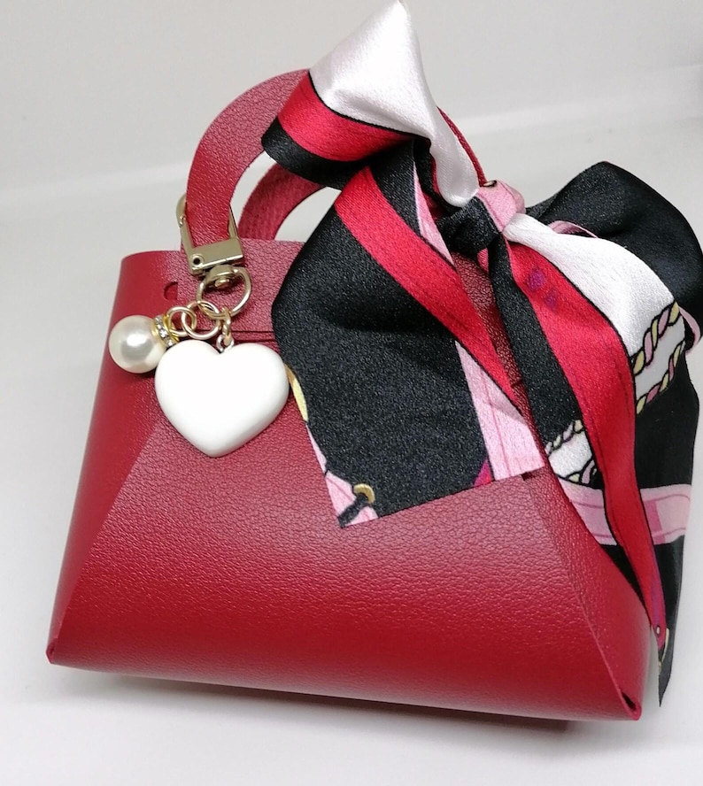 Op de afbeelding: Een kleine, rode handtas met een handvat aan de bovenkant en een decoratieve sjaal die aan de bovenkant is vastgebonden. De tas heeft een wit hartvormig bedeltje en een parelaccent. De sjaal heeft zwarte, rode en witte patronen.