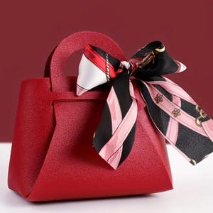Könnte beinhalten: Eine rote Handtasche mit oberem Griff und dekorativem Schal. Der Schal hat ein schwarz-rosa Muster mit floralen Akzenten. Die Tasche hat eine strukturierte Oberfläche und ein strukturiertes Design.