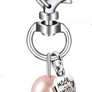 Op de afbeelding: Zilverkleurige sleutelhanger met een hartvormige sluiting. Voorzien van een roze parel engel bedel met zilveren vleugels en een hartvormige hanger met de tekst "Made With Love". Een charmant accessoire.