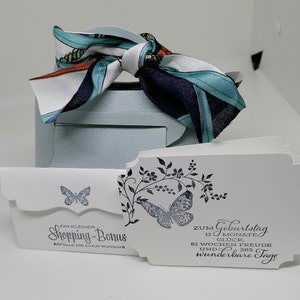 Könnte beinhalten: Hellblaue Geschenkbox mit gemustertem Schal obenauf. Weiße Karten mit Schmetterlings- und Blumendesigns sind vorne. Auf den Karten steht "Shopping-Bonus" und "Zum Geburtstag 12 Monate Glück, 52 Wochen Freude und 365 wunderbare Tage."