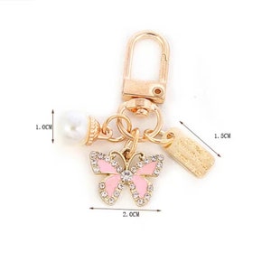 Op de afbeelding: Een goudkleurige sleutelhanger met een roze vlinderbedel, een imitatieparel en een klein rechthoekig label. De vlinderbedel is 2,0 cm breed en versierd met strass. Het label is 1,5 cm lang. De parel heeft een diameter van 1,0 cm.