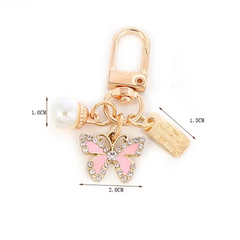 Op de afbeelding: Een goudkleurige sleutelhanger met een roze vlinderbedel, een imitatieparel en een klein rechthoekig label. De vlinderbedel is 2,0 cm breed en versierd met strass. Het label is 1,5 cm lang. De parel heeft een diameter van 1,0 cm.