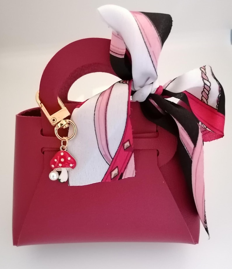 Könnte beinhalten: Burgunderfarbene Handtasche mit goldfarbenem Verschluss und einem Pilz-Anhänger. Ein gemustertes Tuch in Weiß, Rosa und Schwarz ist am Griff gebunden. Der Pilz-Anhänger hat einen weißen Stiel und einen roten Hut mit weißen Punkten.