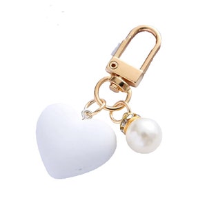 Op de afbeelding: Een witte hartvormige sleutelhanger met een goudkleurige sluiting en accenten. Een imitatieparel met strass accenten bungelt aan de bedel. De sleutelhanger is ontworpen voor sleutels of als decoratief accessoire.