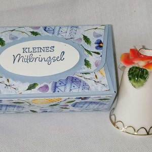 Könnte beinhalten: Eine kleine, hellblaue Geschenkbox mit Blumen- und Eier-Muster und eine weiße Porzellanglocke mit Blumenarrangement. Auf der Box steht der Text "KLEINES Mitbringsel". Die Glocke hat einen goldenen Rand.
