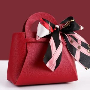 Op de afbeelding: Een kleine, rode handtas met een handvat aan de bovenkant en een decoratieve sjaal aan het handvat. De sjaal heeft een zwart-roze patroon met gouden accenten. De tas heeft een textuuroppervlak en een geometrische vorm.