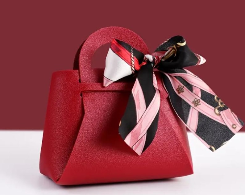 Op de afbeelding: Een kleine, rode handtas met een handvat aan de bovenkant en een decoratieve sjaal aan het handvat. De sjaal heeft een zwart-roze patroon met gouden accenten. De tas heeft een textuuroppervlak en een geometrische vorm.