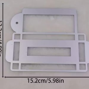 Könnte beinhalten: Eine silberfarbene Metallschablone mit rechteckigem Design. Sie hat einen oberen Abschnitt mit einem kreisförmigen Element und einer rechteckigen Öffnung sowie einen unteren Abschnitt mit einer rechteckigen Öffnung. Die Schablone ist 15,2 cm breit und 12,2 cm hoch.