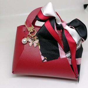 Op de afbeelding: Een kleine, rode handtas met een handvat aan de bovenkant en een decoratieve sjaal aan het handvat. De sjaal heeft zwarte, rode en witte patronen. Een goudkleurige bedel met een parel en roze vlinderaccenten hangt aan het handvat.