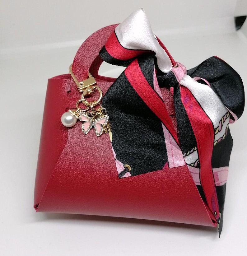 Op de afbeelding: Een kleine, rode handtas met een handvat aan de bovenkant en een decoratieve sjaal aan het handvat. De sjaal heeft zwarte, rode en witte patronen. Een goudkleurige bedel met een parel en roze vlinderaccenten hangt aan het handvat.