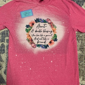 May include: A pink t-shirt with a white bleach design featuring a floral wreath around a hexagon with the text "Aunt A double blessing. You love like a parent And act like a friend."