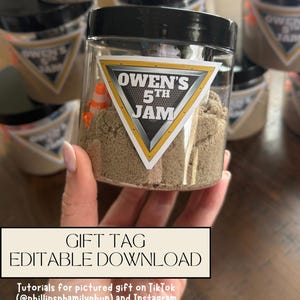 May include: A clear plastic jar with a black lid filled with light brown sand and a small orange and white traffic cone. A triangular label on the front features a metallic border and the text "OWEN'S 5TH JAM" in bold white and black lettering.