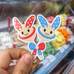 May include: Two cartoon ice cream cone stickers with bunny ears. One is red and white, the other is blue and white. They are holding hands with a red and blue bow.