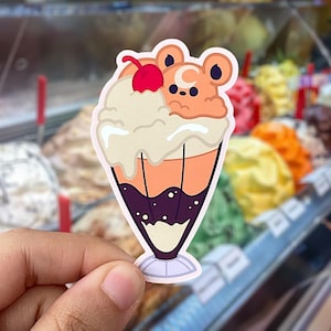 May include: A die-cut sticker depicting a cartoon sundae with a bear face. The sundae has a cherry on top, vanilla ice cream, and a dark purple base. The sticker is held in a hand, with a blurred background of ice cream flavours.