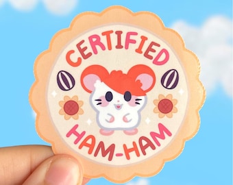 Op de afbeelding: Een ronde sticker met een witte hamster met rood haar, omringd door zonnebloemen en zaden. De sticker zegt "CERTIFIED HAM-HAM" in een roze en oranje lettertype.